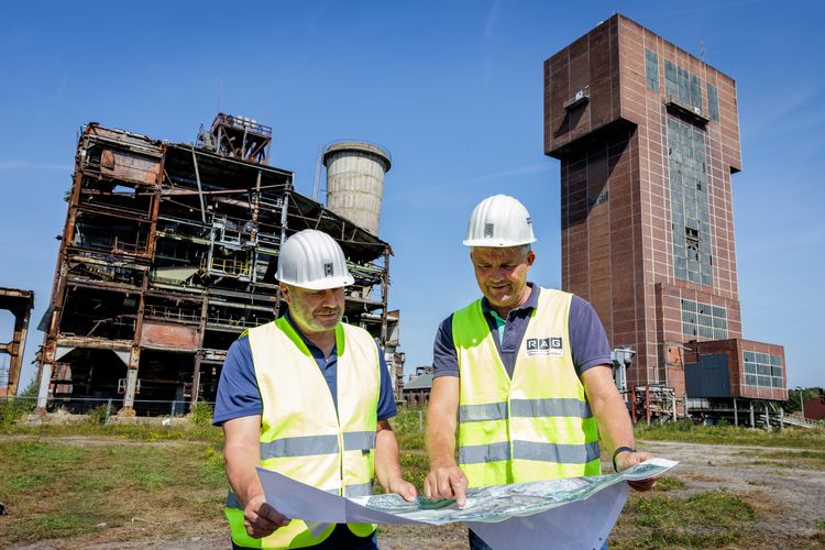 Die zuständigen Projektingenieure der RAG Montan Immobilien, Andreas Boermann und Kai Kopke. Im Hintergrund rechts das Schachtgerüst Robert (Hammerkopfturm), das erhalten bleibt. Links das Schachtgerüst Heinrich mit der Schachthalle und dem Grubenlüfter, die zurückgebaut werden. Foto: RAG Montan Immobilien GmbH/Holger Jacoby